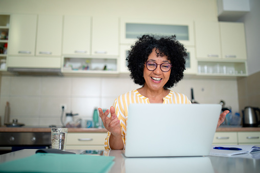Jacqui (woman) smiling during an online GP consultation on a laptop at her kitchen table, in a calm home setting.